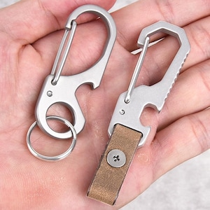 Może przedstawiać: Dwa srebrne metalowe breloki z otwieraczami do butelek. Jeden brelok ma brązowy pasek skórzany i srebrną śrubę. Drugi brelok ma srebrny pierścień.