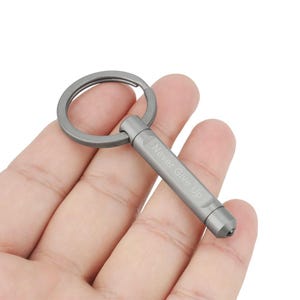 Puede incluir: Un llavero de metal gris con un anillo y un cuerpo cilíndrico. El cuerpo del llavero tiene grabadas las palabras "Never Give Up". El llavero es un accesorio pequeño y compacto.