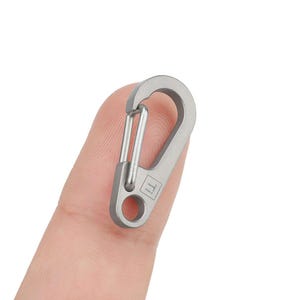 Op de afbeelding: Een zilverkleurige metalen karabijnhaak met een kleine lus aan de zijkant. De karabijn heeft een klein 'T'-logo aan de voorkant.