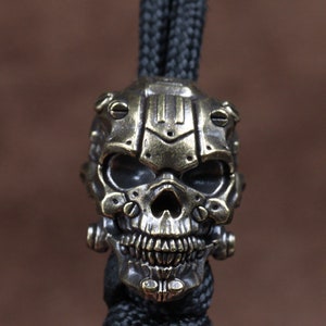 May include: A brass skull bead with a detailed, futuristic design. The skull has a helmet-like structure with screws and lines, and the eyes are hollow and dark. The bead is attached to a black cord.
