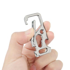 Könnte beinhalten: Ein silberner Multi-Tool-Karabiner mit Haken, Flaschenöffner und anderen integrierten Werkzeugen. Der Karabiner wird in einer Hand vor weißem Hintergrund gehalten. Das Werkzeug hat eine Vielzahl von Aussparungen und ein robustes Design.