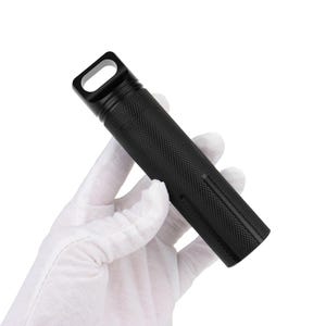 Può includere: Un contenitore cilindrico nero con una superficie strutturata e un anello rettangolare nella parte superiore. Il contenitore è tenuto in una mano guantata su sfondo bianco. Il contenitore ha un design a coste.