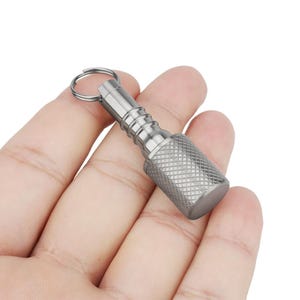 May include: A small, silver-colored keychain accessory with a textured grip and a ring for attaching keys. The cylindrical body has a series of ridges and a flat end. The item is held in a hand.