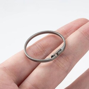 May include: A silver-colored ring with a unique clasp design. The ring is circular with a small rectangular clasp that appears to lock the ring together. The ring is held in a person's hand.