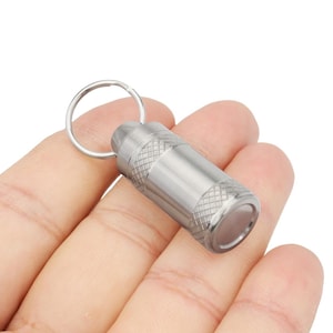May include: A small, silver-colored metal keychain pill holder. The cylindrical container has a textured, diamond-patterned grip on the ends and a smooth, polished center. A silver key ring is attached to the top.