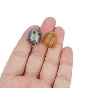 Könnte beinhalten: Zwei geisterförmige Flaschenverschlüsse, einer silberfarben und einer golden, mit einem Loch in der Mitte jedes. Die Verschlüsse liegen auf einer Hand.