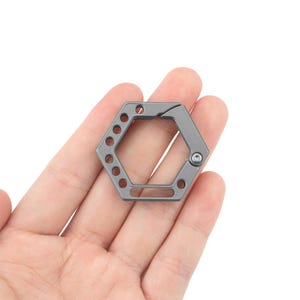 May include: A silver hexagonal key chain with a small hole on the side. The key chain is designed to hold keys and other small items.