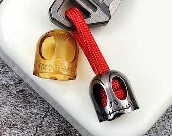 May include: Two skull-shaped keychains, one is amber coloured plastic and the other is silver metal. Both keychains have a stitched mouth and eyes. The keychains are attached to a red cord.