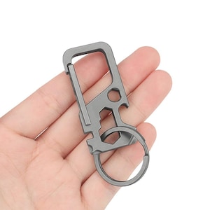 Puede incluir: Un llavero multiherramienta gris con un mosquetón, un abrebotellas y un llavero. La herramienta de metal se sostiene en una mano sobre un fondo blanco. La herramienta tiene una abertura hexagonal y un destornillador plano.