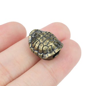 Peut inclure: Un petit charme de tortue en métal doré, détaillé. La tortue est tournée vers le bas, sa carapace est visible. La carapace a un design texturé et écailleux.