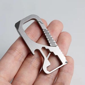 May include: A silver multi-tool with a carabiner clip, bottle opener, and other integrated tools. The tool has a textured grip and a pointed edge. The design is compact and appears to be made of metal.