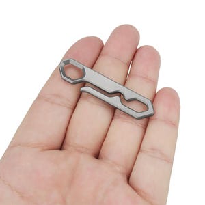 Op de afbeelding: Een kleine, zilverkleurige multitool met een zeshoekige opening en een gebogen haak. De tool wordt in een hand gehouden, wat de compacte afmetingen en het ontwerp benadrukt. De tool lijkt van metaal te zijn en heeft een minimalistische uitstraling.