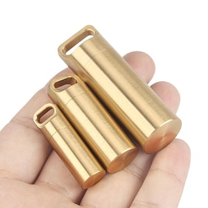 May include: Three cylindrical brass containers of varying sizes, each with a loop at the top. The containers are held in a hand, showcasing their compact design and metallic sheen. The smallest container is approximately 1 inch in length.