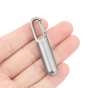May include: A small, silver capsule with a carabiner clip. The capsule has a rounded base and a smooth, metallic finish. The clip is attached to the top, allowing it to be easily attached to a keyring or bag. The capsule is approximately 6 cm in length.