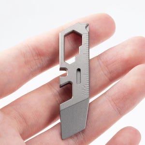 Puede incluir: Una pequeña multiherramienta plateada con una regla integrada marcada en centímetros. La herramienta presenta una abertura hexagonal, un abrebotellas y una pequeña cuchilla. El diseño es compacto y parece ser de metal.