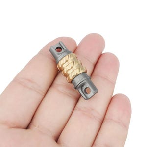 May include: A small, silver and gold metal fidget toy with a textured, grooved design. The toy has two holes on one end and a single hole on the other end.