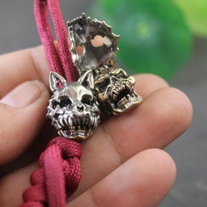 May include: A close-up shot of a metal wolf head and skull bead set on a red cord. The wolf head is silver, and the skull is bronze. The beads have intricate details and are held in a hand.
