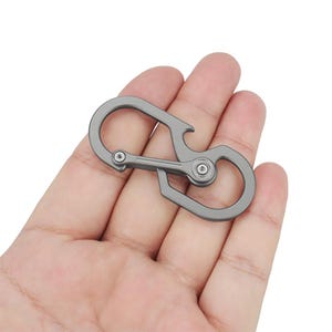 May include: A silver metal carabiner with a bottle opener attached. The carabiner has a smooth, rounded shape and a spring-loaded gate.