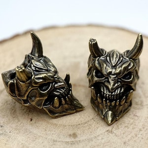 May include: Two detailed, ornate, bronze-colored, metal, oni mask charms. The masks have sharp, pointed horns, fierce expressions, and intricate details.
