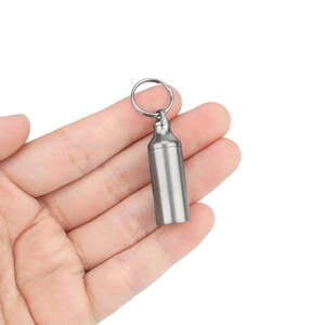 May include: A silver metal keychain pill container with a screw-on lid. The container is attached to a silver metal key ring.