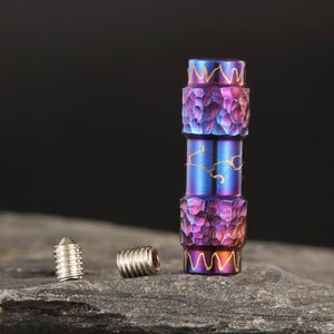 May include: A multi-colored metal bead with a textured surface and gold accents. The bead is cylindrical with a series of stacked sections. Two small silver screws are next to the bead. The bead has a purple, blue, and gold color scheme.
