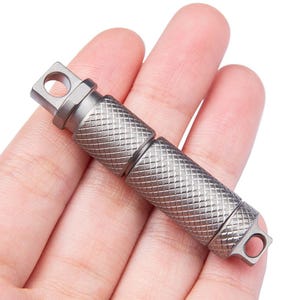 Könnte beinhalten: Ein silberner Schlüsselanhänger aus Metall mit einer strukturierten Oberfläche und einer kleinen Schlaufe an jedem Ende. Der Schlüsselanhänger ist als Multitool konzipiert.