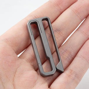 May include: A gray metal money clip with a rectangular design. The clip has a unique shape with a central bar and a hinged section for securing bills. The clip is shown in a hand.
