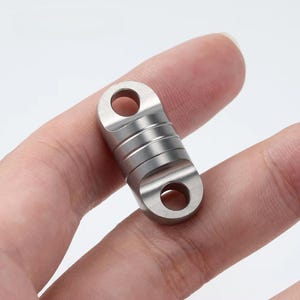 May include: A small, silver-colored metal object with two circular holes at each end and a ridged, cylindrical center. The object is held in a person's hand, showcasing its compact size and design.