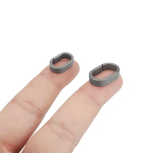 Könnte beinhalten: Zwei kleine, oval geformte, dunkelgraue Metallverschlüsse. Jeder Verschluss ist auf einer Seite offen. Die Verschlüsse werden auf den Fingern einer Person vor weißem Hintergrund gezeigt. Diese Verschlüsse werden wahrscheinlich für die Schmuckherstellung oder andere Bastelprojekte verwendet.