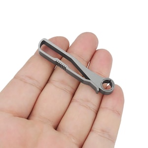 May include: A silver titanium keychain with a small loop on one end and a larger loop on the other end. The keychain has a textured surface and a small groove in the middle.