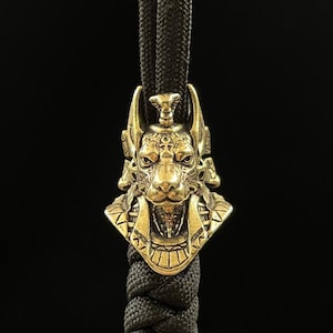 Puede incluir: Un amuleto de metal dorado que presenta una representación detallada de una cabeza de Anubis con una cobra en la parte superior. El amuleto está unido a un cordón negro.