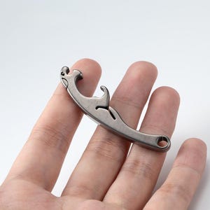 May include: A silver-colored bottle opener with a curved design. The opener has a hook-like feature and a small hole at the end. The object is held in a person's hand against a white background.