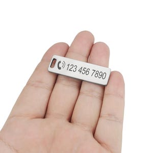 Könnte beinhalten: Ein rechteckiger, silberfarbener Tieranhänger mit abgerundeten Ecken. Der Anhänger zeigt ein Telefonsymbol und die Telefonnummer "123 456 7890" in einer einfachen, serifenlosen Schrift eingraviert. Der Anhänger hat ein kleines Loch zur Befestigung.