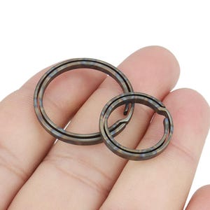 Puede incluir: Dos llaveros metálicos en una mano. Los anillos son de color bronce oscuro con detalles azules y negros. Un anillo es más grande que el otro. Los anillos son circulares con una abertura dividida.