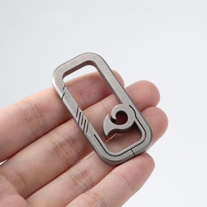 May include: A rectangular, silver-colored carabiner with a unique hook design. The carabiner features a series of diagonal lines on one side and a curved hook on the other. The metal has a matte finish.