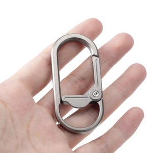 Könnte beinhalten: Ein silberfarbener Karabinerhaken mit ovaler Form und einem Scharnierverschluss. Der Haken wird in einer Hand vor weißem Hintergrund gehalten. Er hat ein kleines Schraubendetail in der Nähe des Scharniers.