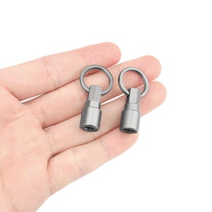 May include: Two silver swivel key rings with a small metal loop at the top and a threaded base. The key rings are held in a hand.