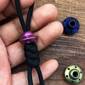 Puede incluir: Una cuenta de color morado con forma de OVNI con un cordón de paracord negro. La cuenta tiene una superficie texturizada y está adornada con un pequeño agujero en el centro. Se muestran dos cuentas adicionales en el fondo, una azul y otra verde.