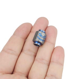 Pode incluir: Uma pequena conta de titânio cinza com um design de caveira sorridente em esmalte azul. A conta é projetada para ser usada com um bracelete de paracord.