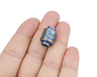 May include: A small, grey titanium bead with a blue enamel design of a smiling skull. The bead is designed for use with a paracord bracelet.