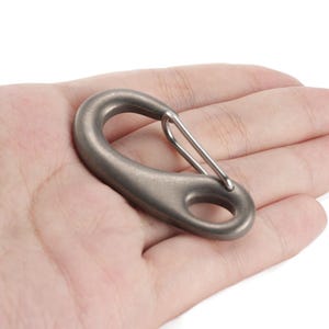 Può includere: Un moschettone grigio metallizzato con un cancello a molla. Il moschettone ha una forma ovale con un corpo arrotondato e un piccolo foro nella parte inferiore. Il moschettone è tenuto in una mano.