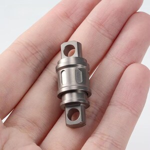 Peut inclure: Un petit composant pivotant métallique avec un corps cylindrique et des extrémités hexagonales, chacune avec une ouverture circulaire. Le composant a une finition en métal brossé et est tenu dans une main, mettant en valeur sa taille compacte et son design.