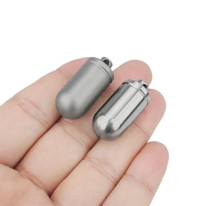 Può includere: Due piccoli contenitori metallici grigi a forma di capsula con un anello nella parte superiore. Uno ha una finitura opaca, l'altro è lucido. I contenitori sono tenuti in una mano, suggerendo una piccola dimensione, forse per trasportare pillole o piccoli oggetti.