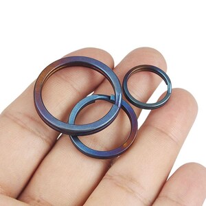 May include: Three iridescent metal key rings in shades of blue, purple, and gold are displayed. The rings are circular, with one larger ring interlocked with a smaller ring. The smallest ring is separate. The key rings are held in a hand.