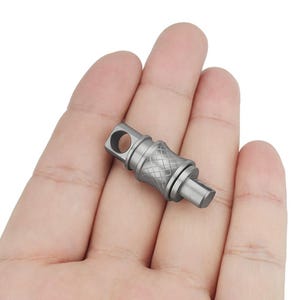 Puede incluir: Un llavero de metal de color plateado con un diseño texturizado y un pequeño lazo. El llavero tiene un pequeño agujero en un extremo y una forma rectangular plana en el otro extremo.