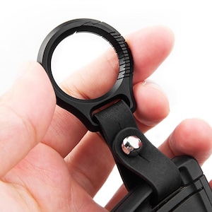 Puede incluir: Una correa de cuero negra con un anillo de metal y un bucle de plástico negro. El bucle tiene una superficie texturizada y un pequeño remache metálico.