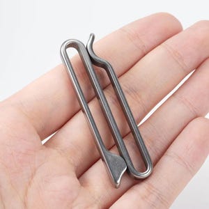Könnte beinhalten: Ein silberfarbener, minimalistischer Geldclip. Der Clip besteht aus Metall und hat ein schlichtes, stromlinienförmiges Design. Er hat eine gebogene Oberseite und eine flache, abgewinkelte Unterseite. Der Clip wird in der Hand einer Person gehalten.