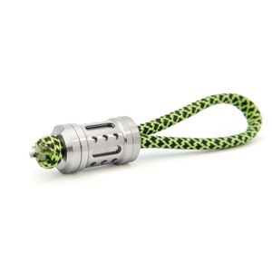 May include: A silver metal key chain with a green and black braided cord. The metal piece has a cylindrical shape with a series of holes.