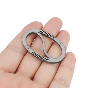 Puede incluir: Un llavero de titanio plateado con un diseño de doble bucle. El llavero tiene una superficie texturizada y se sostiene en una mano.