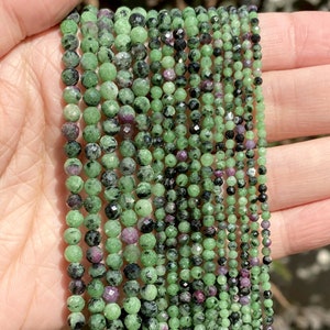 May include: A strand of green, black, and pink gemstone beads. The beads are round and faceted. The strand is approximately 16 inches long.